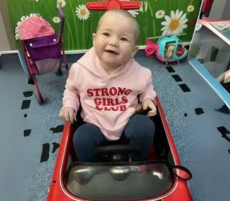 A young girl with little hair sits in a toy car, wearing a pink jumper that says 'Strong Girls Club.'