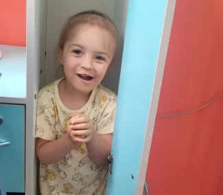 A little girl wearing a yellow t-shirt is playing, hiding inside a blue and red cupboard with the door open, smiling and holding her hands together.