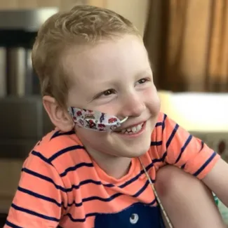 A young boy with a nasogastric tube is smiling while wearing a striped orange t-shirt.