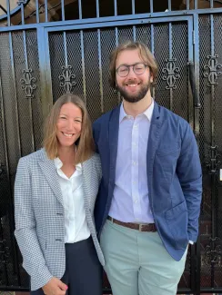 A man with brown hair and glasses stands next to a woman with shoulder-length brown hair, wearing a grey blazer. 