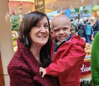 A smiling woman with dark hair is holding her young son, who is bald and wearing a bright red jacket.
