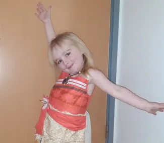 A smiling young girl with blonde hair stands with her arms outstretched, wearing an orange top and a beige skirt.