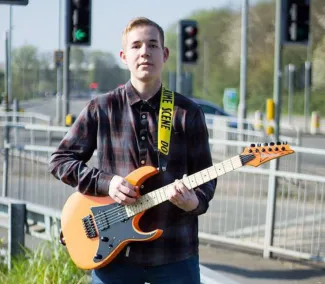A young man is standing next to a pedestrian crossing, holding a guitar.