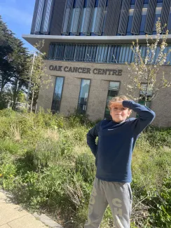 A young boy with curly blonde hair is happily standing outside the Oak Cancer Centre.