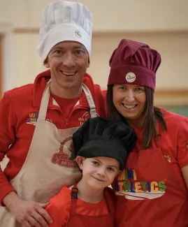 A man, a woman, and a young boy wearing bright red 'Mini Athletics' clothing and chef hats.