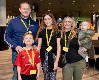 A family of five, including a dad, mum, young daughter, little boy, and baby, standing together wearing yellow lanyards.