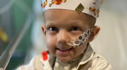 A photo of a smiling young boy sitting on a hospital bed with a tube in his nose, wearing a white paper crown decorated with shiny bits of paper.