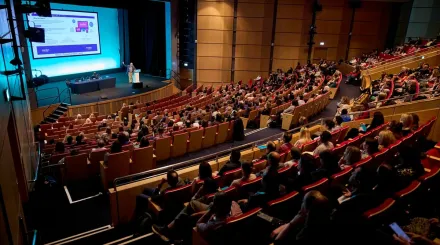 People sitting in a large room, watching someone give a presentation on a stage.