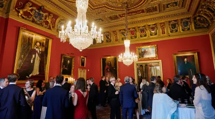An ornate event room with large glass chandeliers and paintings, with people standing around talking in black tie attire