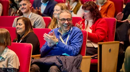 A group of people at a conference, many of them smiling. In the middle is a man smiling and clapping.