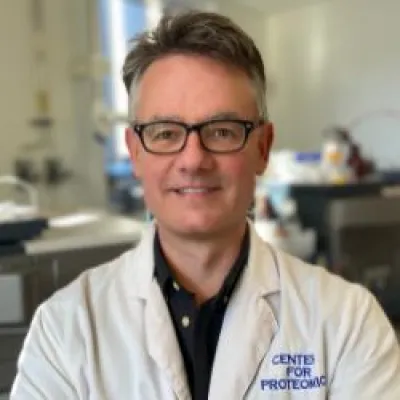 Rob with glasses and short grey hair, smiling at the camera and wearing a white lab coat.