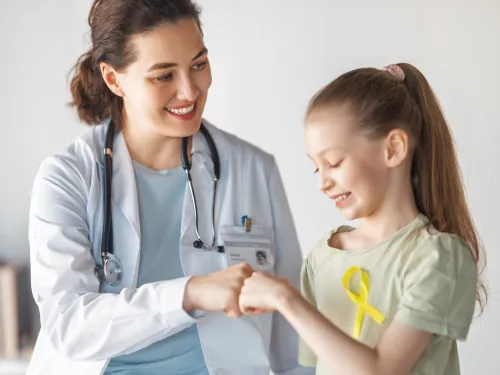 A smiling female doctor interacting with a happy young girl who is wearing a gold ribbon for childhood cancer awareness.