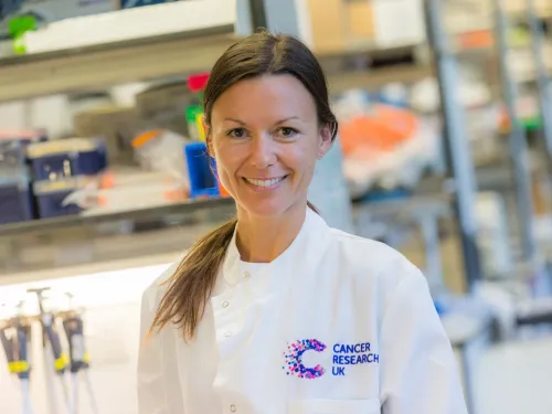 Alejandra in the lab, smiling at the camera and wearing a white lab coat with her brown hair in a ponytail.