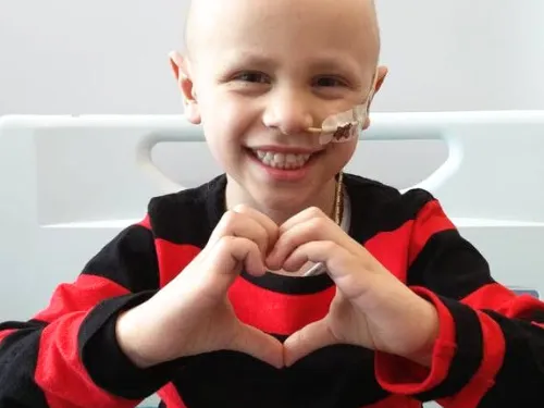 A boy called Ben, who has a feeding tube and has lost his hair, wearing a black and red striped jumper. He is smiling and holding his hands in a heart shape.