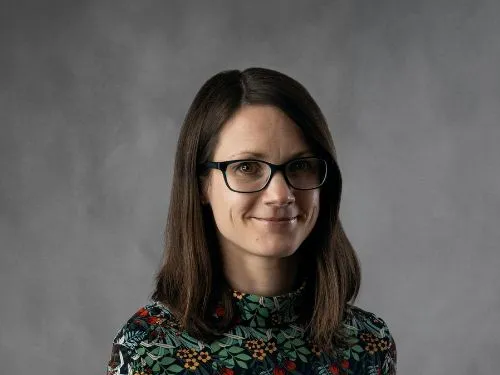 Nicola smiling at the camera. She has dark shoulder length hair, and is wearing a floral blouse.