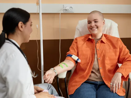 A teenager with a shaved head and orange jacket smiling a a doctor whilst receiving intravenous medicines.