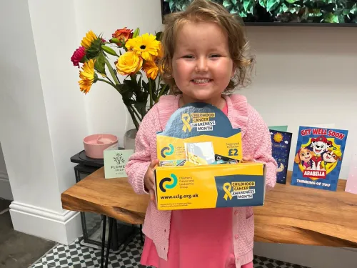 Arabella, a young girl who was diagnosed with acute lymphoblastic leukaemia, smiles at the camera whilst holding a CCLG Childhood Cancer Awareness Month appeal box.