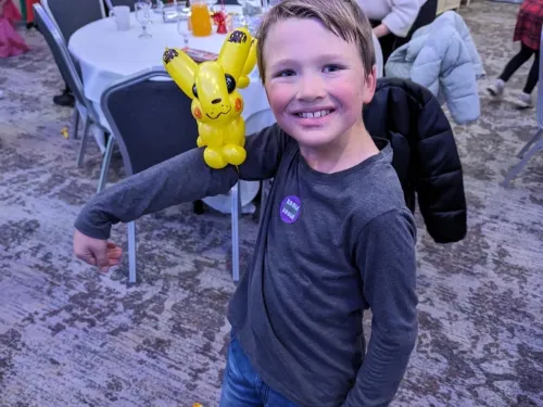 A young boy in a long-sleeve navy top is smiling at the camera with his arm out balancing a yellow balloon character.