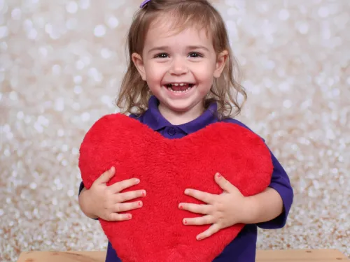 A young girl with brown hair smiles at the camera, wearing a purple top and holding a large red fluffy heart.