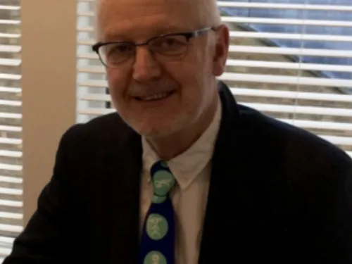 A man with grey hair and glasses is dressed in a white shirt and a black blazer. He is wearing a blue tie with lighter blue circles.