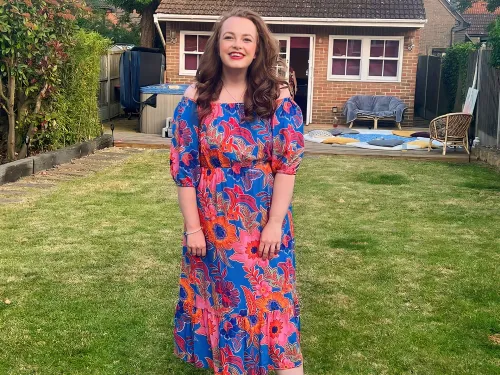 A young woman in her 20s with long brown hair is standing in a garden, smiling at the camera. She is wearing a red and blue floral dress.