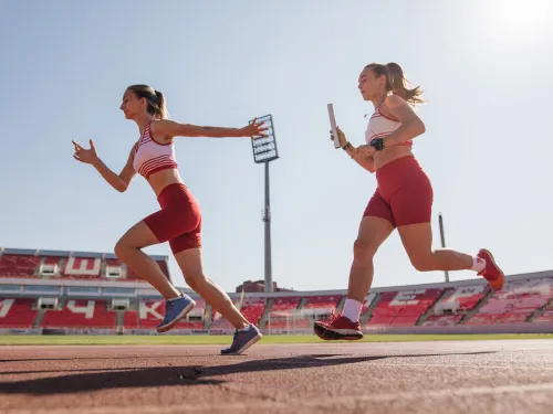 Two women running a relay race in red shorts and white crop tops.