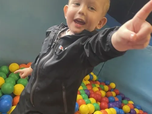 A young boy with short blonde hair is playing in a colourful ball pit, smiling and pointing past the camera.