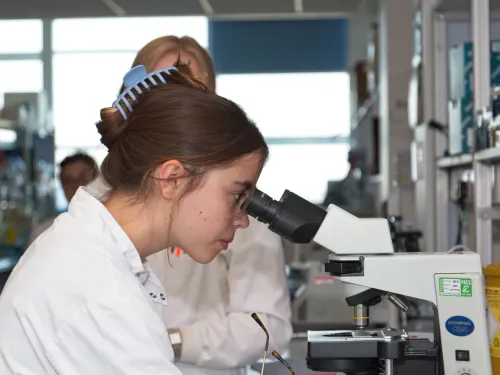 Tabitha, with her brown hair in a claw clip, wearing a lab coat and looking into a microscope.