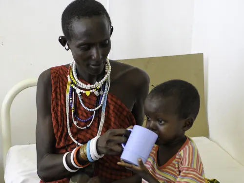 A mother in traditional Tanzanian clothes helping her son to drink whilst sat on a hospital bed.