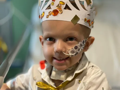 A photo of a smiling young boy sitting on a hospital bed with a tube in his nose, wearing a white paper crown decorated with shiny bits of paper.