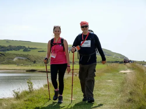 A man and woman in walking gear are smiling as they hike past a lake on a sunny day