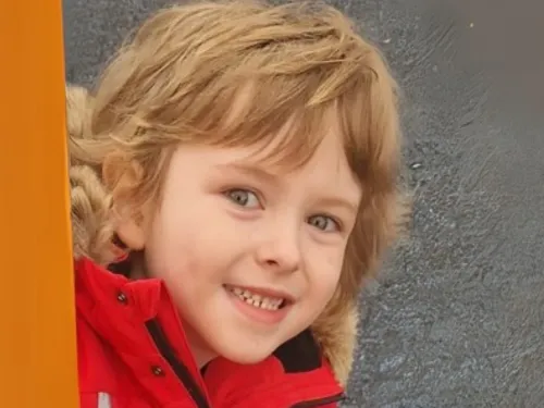 A young boy smiles at the camera while peeking around an orange climbing frame.