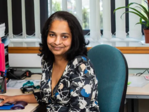 Madhu sat at a desk and smiling at the camera. She is wearing a printed dress and has her dark hair down.