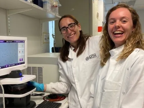 Maria with PhD student Charlie. They are both wearing lab coats and smiling at the camera, stood in front of lab equipment.