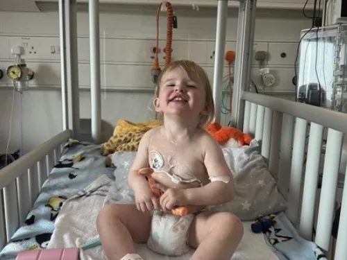 A young girl wearing a nappy is sitting upright in a hospital cot, surrounded by various medical equipment.