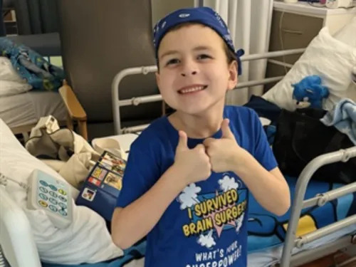 A young boy wearing a blue hat and t-shirt is leaning against a hospital bed, giving a thumbs up to the camera.