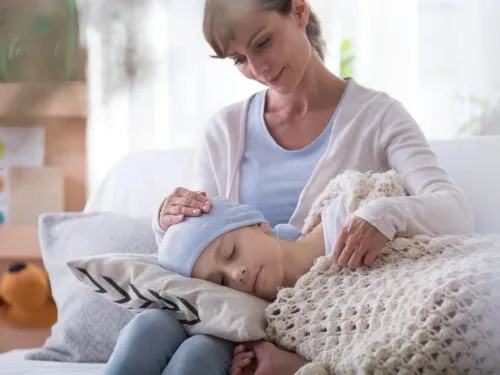 A mother sits on the sofa, with her sick child's head on a cushion in her lap.