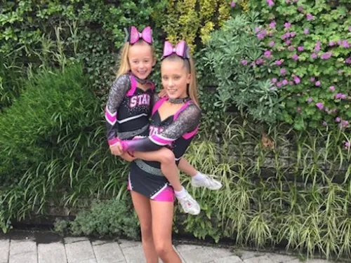 Two girls stand together in pink and black cheerleading uniforms. The older girl is holding the younger girl up.