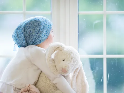 Child with cancer looking out of the window whilst hugging a stuffed rabbit toy.