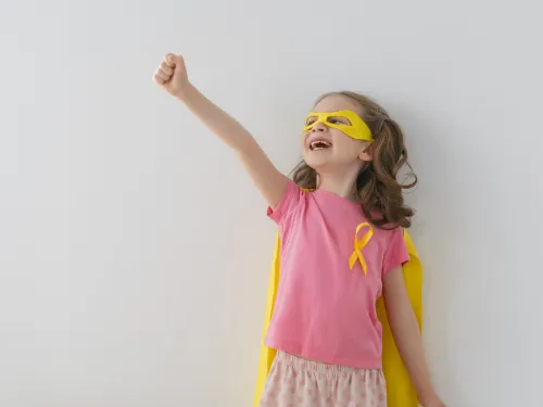 A girl in a pink t-shirt and yellow cape, wearing a gold ribbon badge and posing like a superhero.