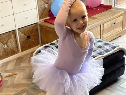 A young girl with a bald head is wearing a purple tutu, dancing like a ballerina.