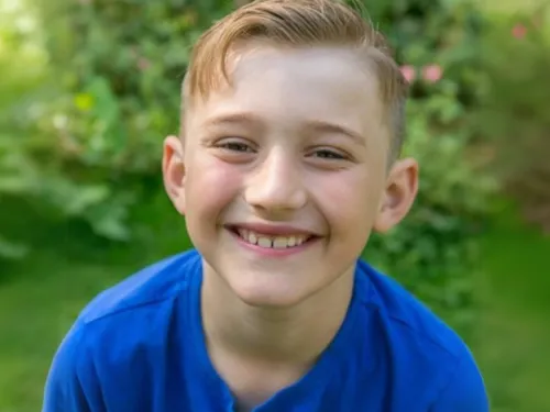 A smiling young boy with short blonde hair is standing in a sunlit garden, wearing a bright blue t-shirt.