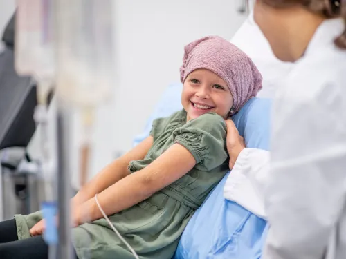 A young girl in hospital, smiling and wearing a pink headscarf whilst talking to a doctor.