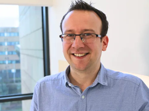 Tim wearing glasses and smiling at the camera. He has short brown hair and is wearing a pale blue shirt.