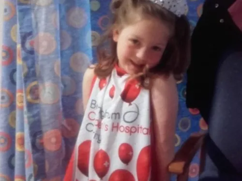 A young girl with long brown hair in pigtails is smiling in front of a hospital curtain, wearing a Birmingham Children's Hospital gown. 