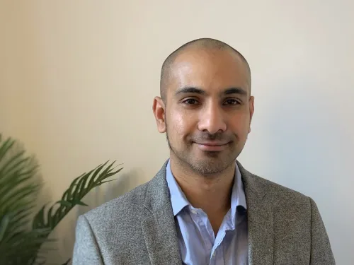 Ruman Rahman smiling at the camera. He has a shaved head and is wearing a grey blazer with a pale blue shirt.