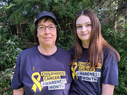  A mother and daughter stand close together, wearing matching navy blue t-shirts with a Childhood Cancer Awareness Month logo on.