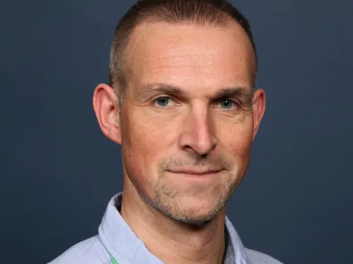 Gareth smiling at the camera in front of a plain blue backdrop. He has very short hair and is wearing a blue shirt.