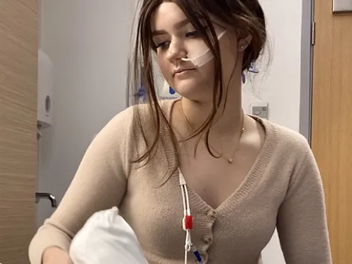 A young girl with brown hair tied back sits in a hospital bed. She has a nasogastric tube inserted and is wearing a beige cardigan.