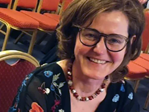 Maureen smiling at the camera whilst sat on a red chair. She has dark hair in a cob and is wearing glasses and a floral dress.